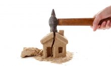 Eine Hand schlägt mit einem Hammer auf ein Haus aus Sand und zerstört es | Notverkauf Immobilie