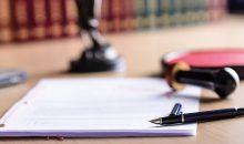 Auf einem Tisch liegen ein Kaufvertrag, ein Füller und ein Stempel, im Hintergrund stehen Rechtsbücher | Notartermin